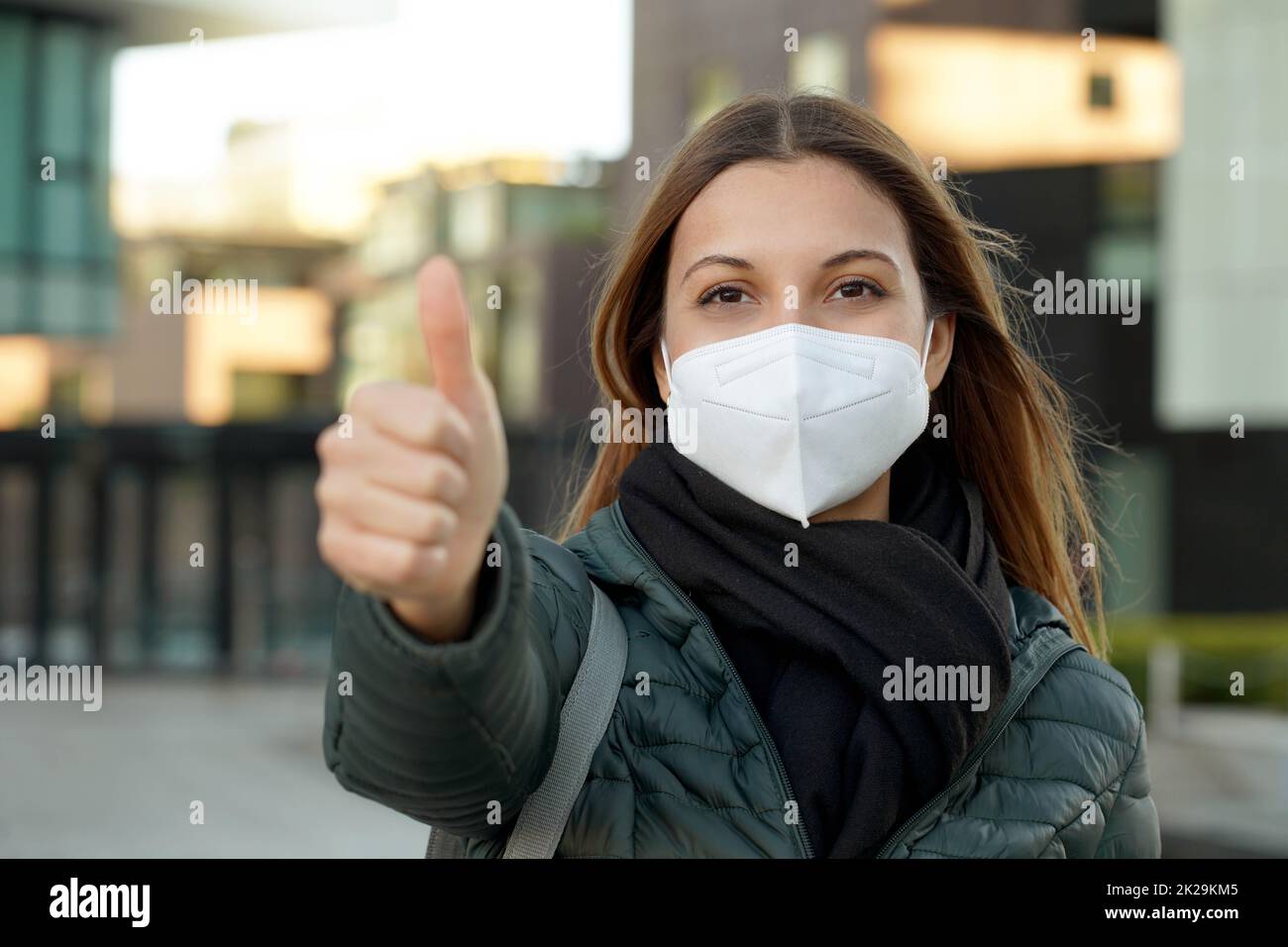 Giovane studentessa ottimista che dà i pollici in su il gesto. Indossa una maschera protettiva (DPI) per evitare l'inquinamento atmosferico o la pandemia di coronavirus per motivi di sicurezza. Foto Stock