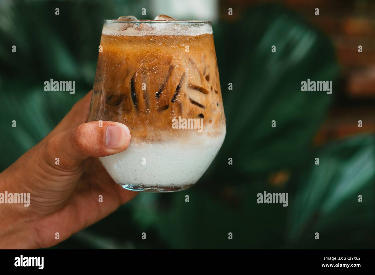 Tenere a mano il caffè latte ghiacciato in un bicchiere con la crema sul fondo. Freddo fondo bevanda estiva Foto Stock