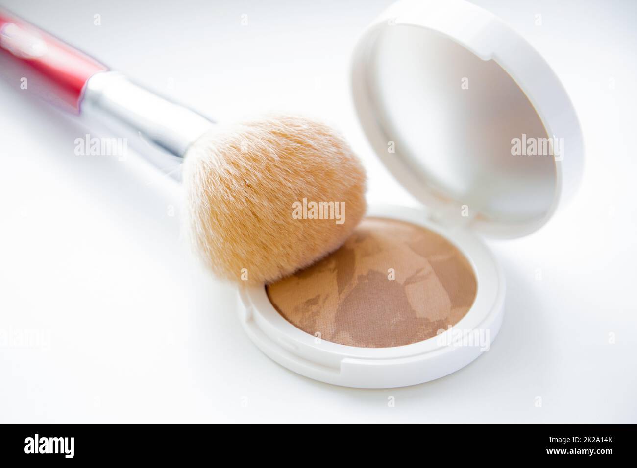 Un pillbox bianco con polvere e bronzer giace su uno sfondo bianco, su di esso giace una spazzola per arrossire in cima. Foto Stock