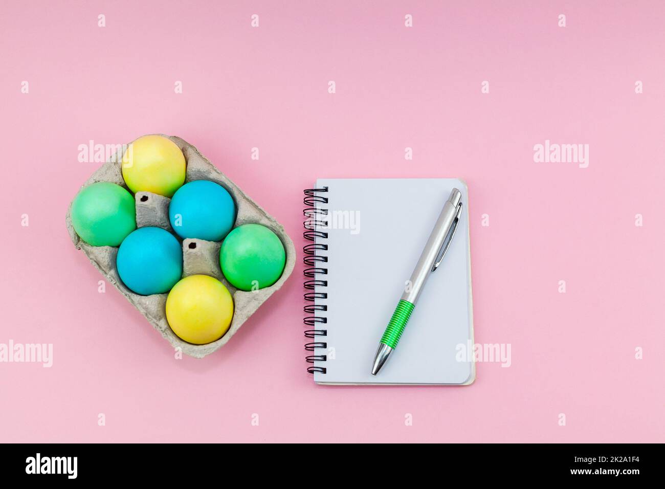 Blocco note vuoto e penna su sfondo rosa scuro alla moda. Vassoio con uova di Pasqua multicolore vicino al notebook. Foto Stock