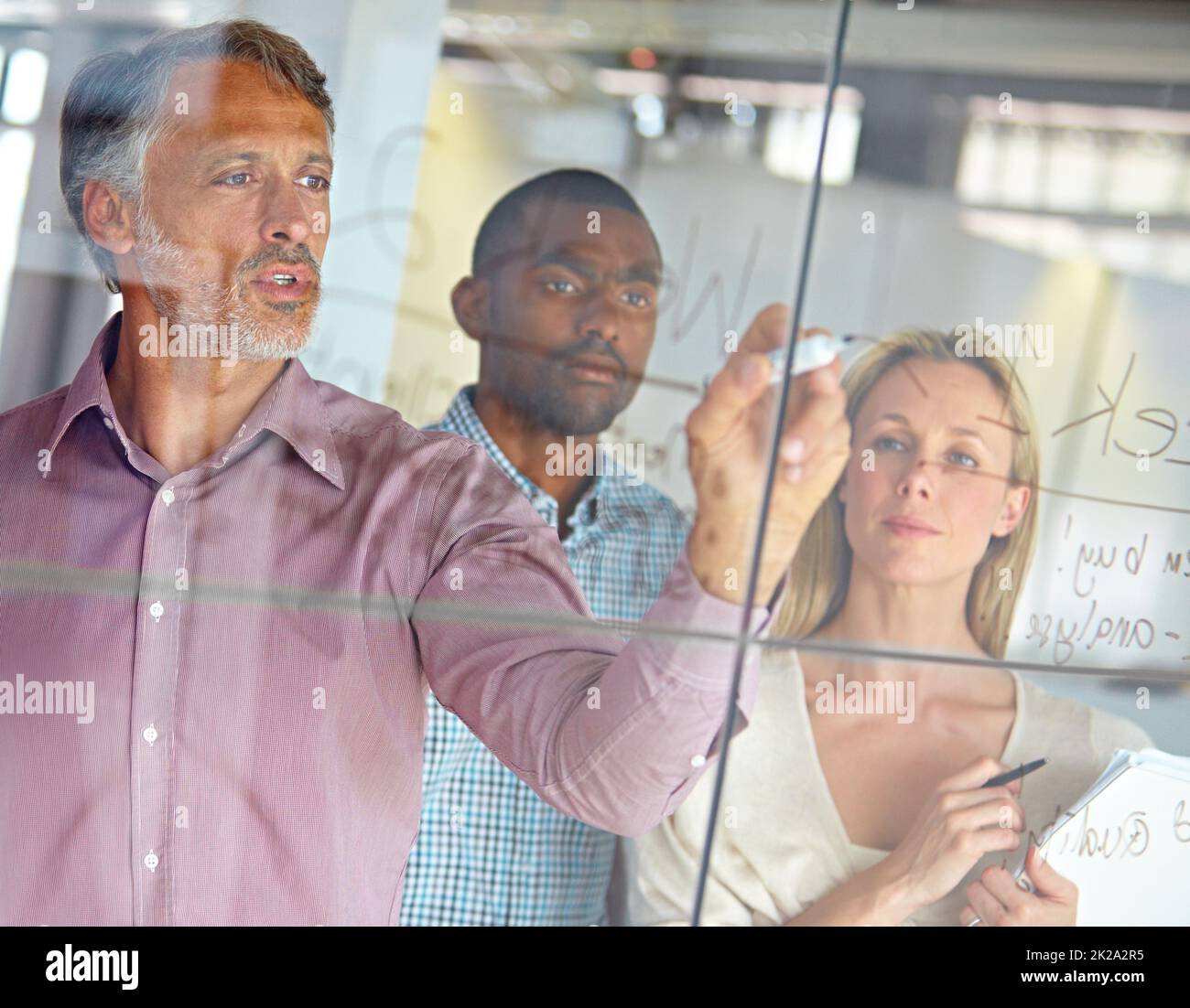 HES un uomo con un piano. Un uomo d'affari maturo che scrive i programmi su un vetro mentre i suoi colleghi guardano sopra. Foto Stock