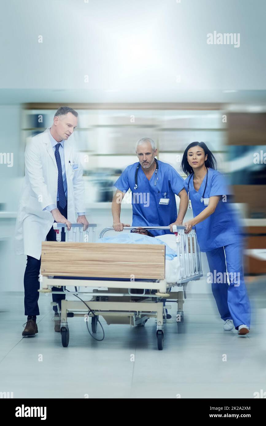 Alla stat sala operatoria. Shot di un gruppo di professionisti medici che precipitano un paziente su un gurney lungo un corridoio ospedaliero. Foto Stock