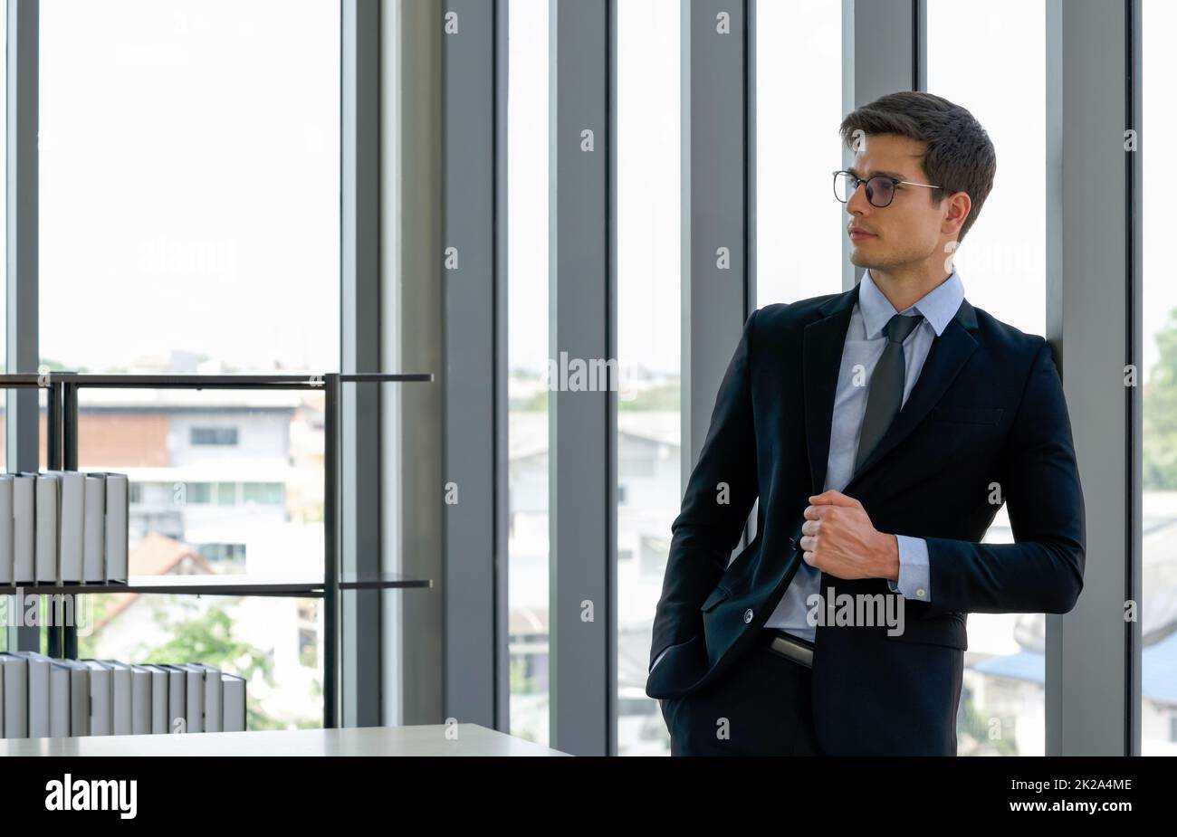 Il giovane uomo d'affari caucasico ha messo la mano nel pant della tasca mentre tiene il taglio anteriore del vestito nero, appoggiandosi contro una parete di vetro nell'ufficio. Atmosfera di lavoro mattutina in un ufficio moderno. Foto Stock