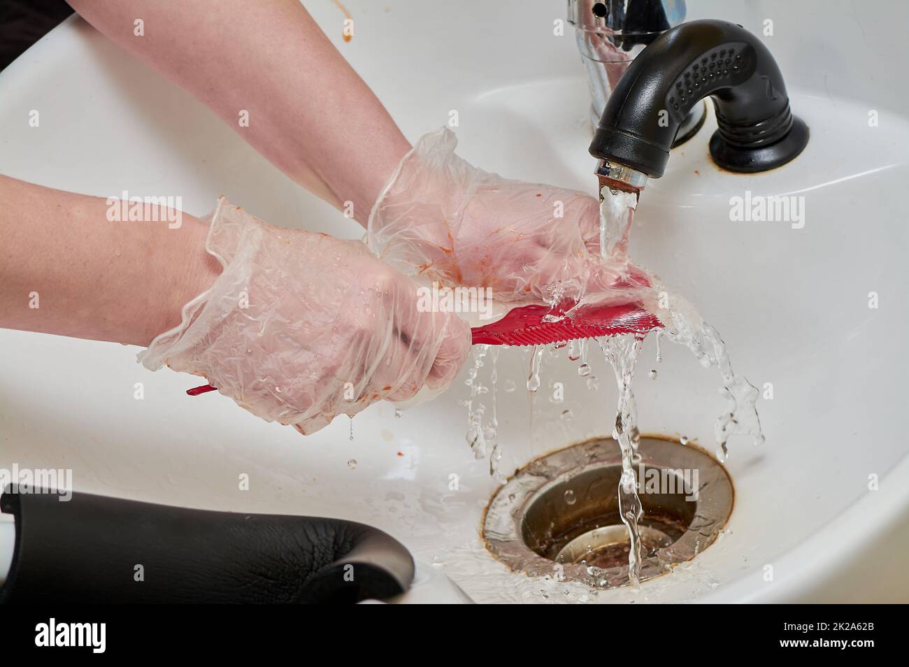 Le mani delle donne nei guanti da colorare dei capelli lavano un pettine sotto un flusso d'acqua in un lavandino bianco Foto Stock