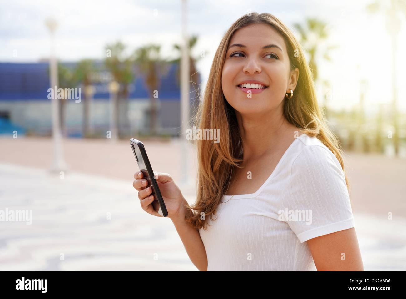 Ragazza sorridente che passeggava da sola tenendo il telefono al tramonto in estate Foto Stock