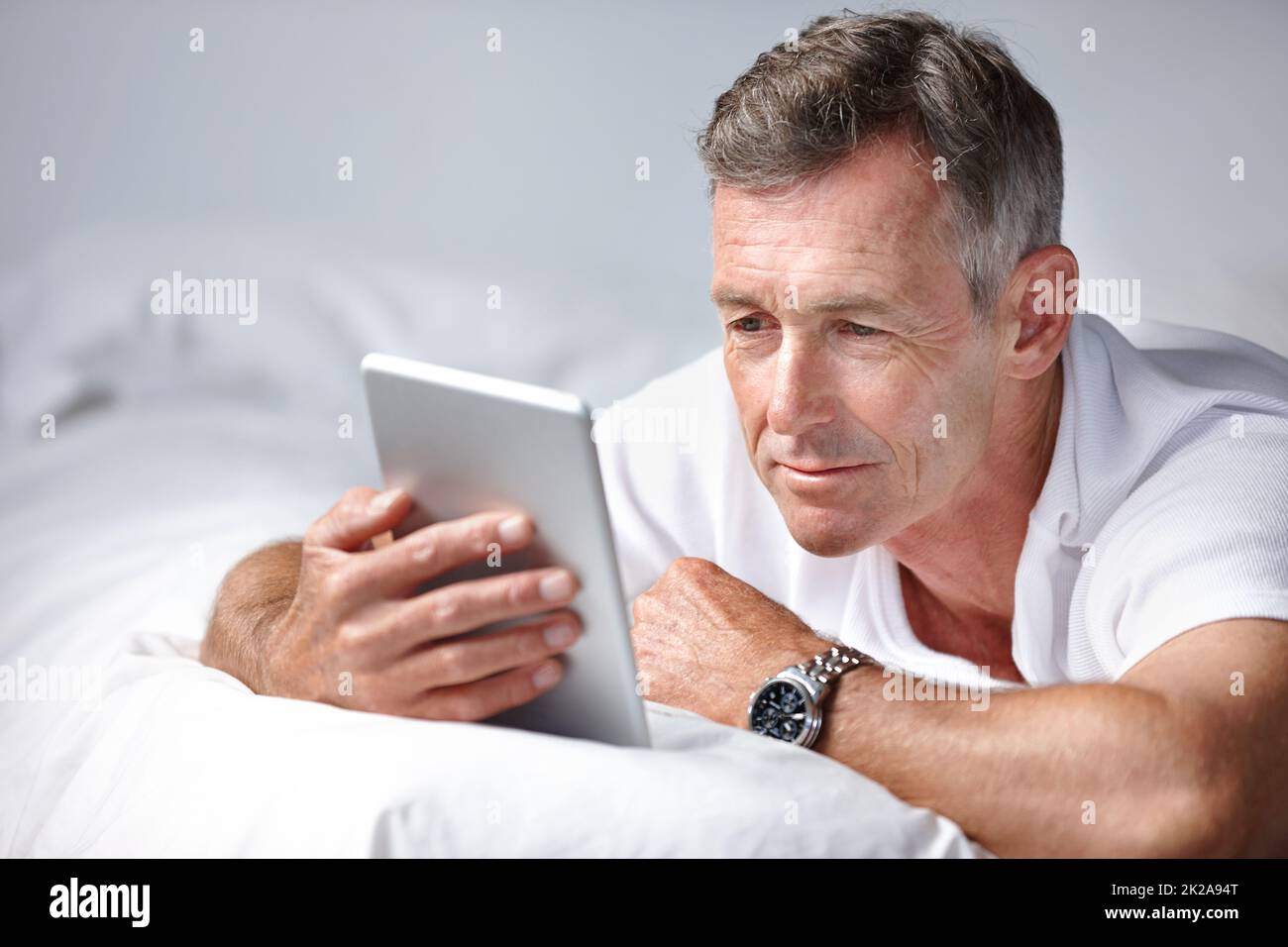 Leggere un interessante articolo online. Scatto di un uomo maturo usando un tablet digitale mentre si trova sul suo letto. Foto Stock