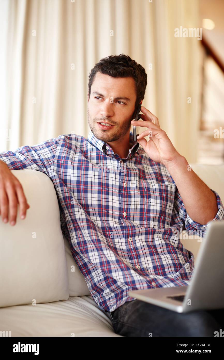 Lavorare da casa. Un bell'uomo che parla sul suo cellulare mentre si siede con il suo laptop sul divano. Foto Stock