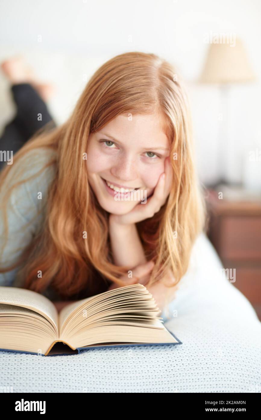 Godersi il mondo della fiction. Ritratto di una giovane redhead che legge il suo libro e si trova sul suo letto. Foto Stock