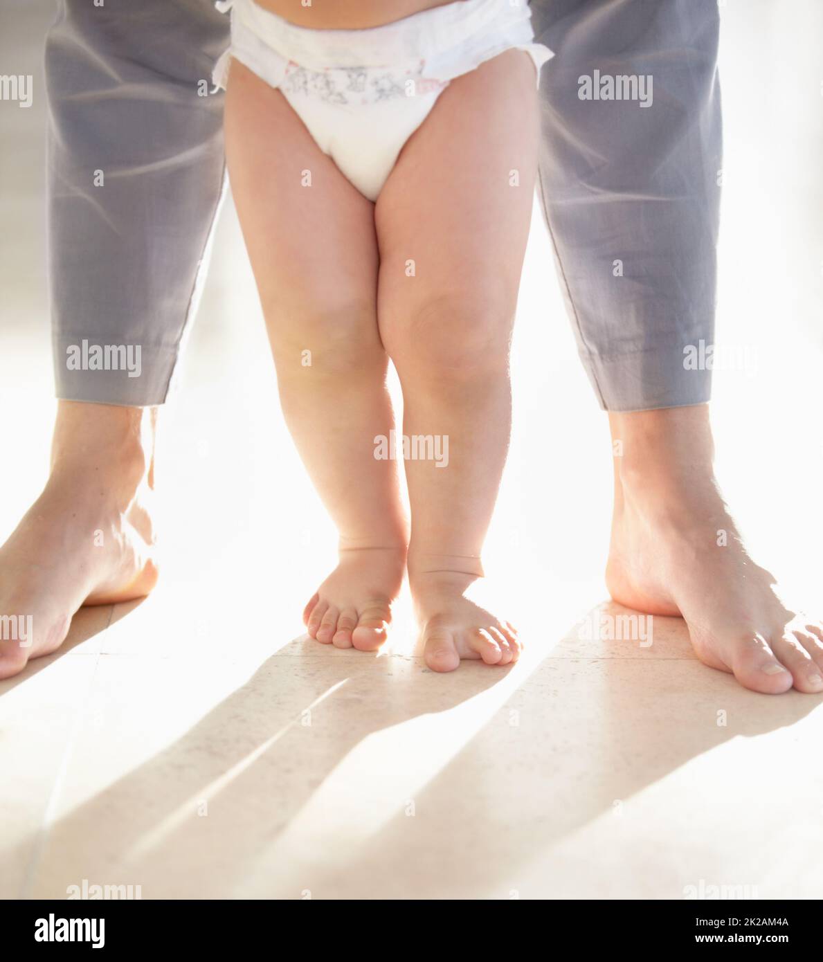 Primi passi. Immagine ritagliata delle gambe di un bambino che cammina con un genitore che sostiene da dietro. Foto Stock