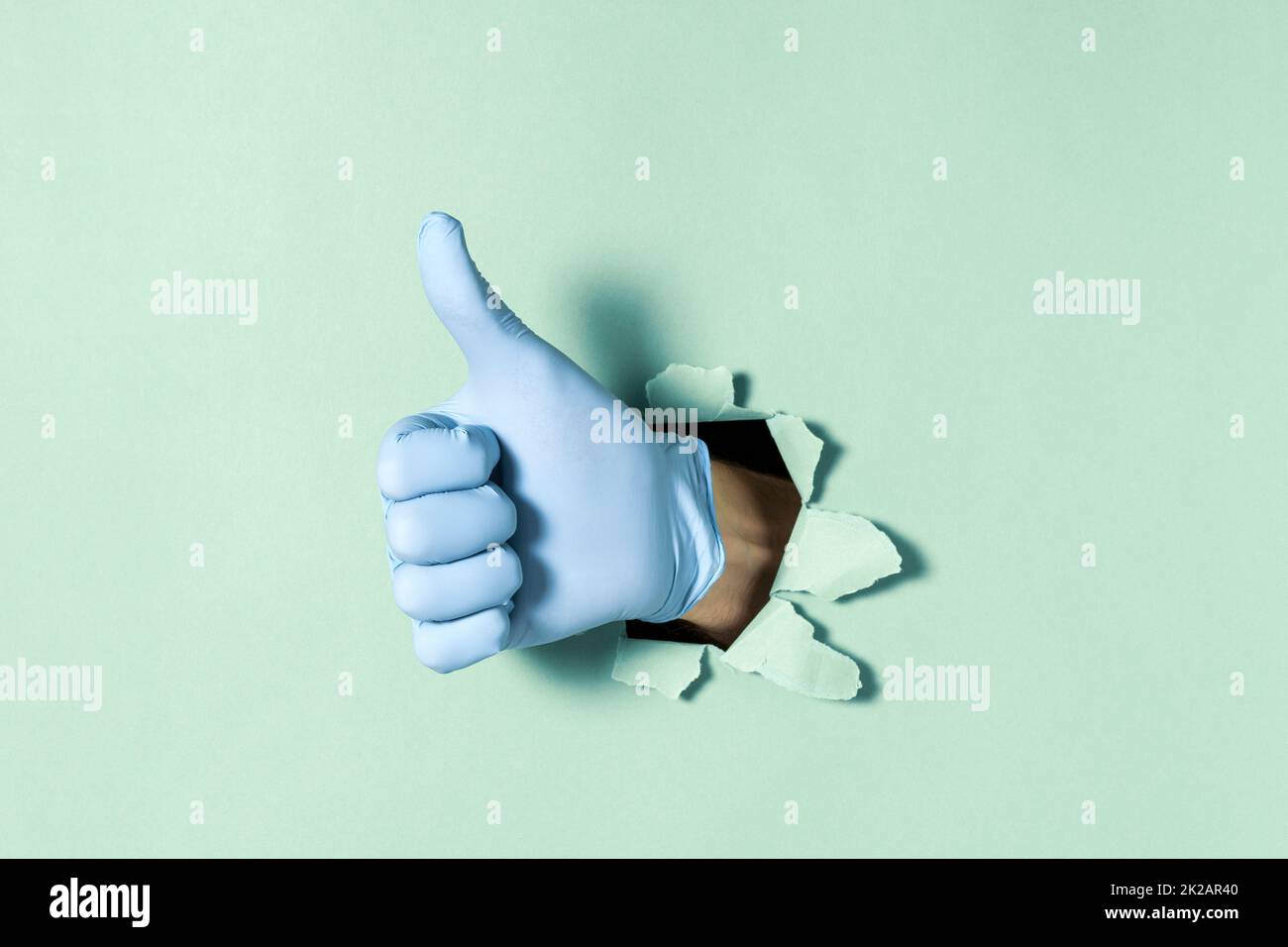 Mano in guanto medico con il pollice verso l'alto gesto attraverso il foro nella parete di carta Foto Stock