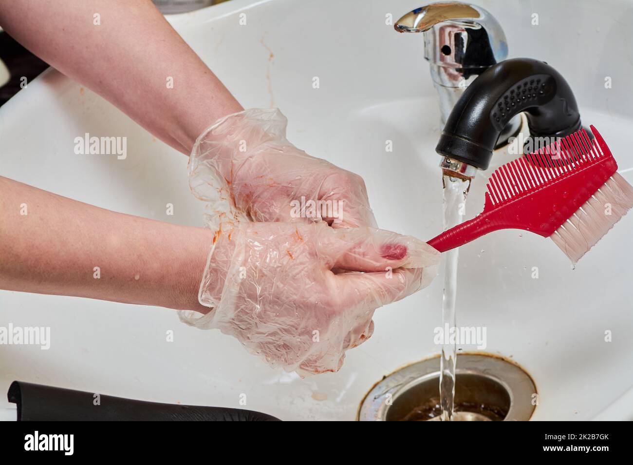 Le mani delle donne nei guanti da colorare dei capelli lavano un pettine sotto un flusso d'acqua in un lavandino bianco Foto Stock