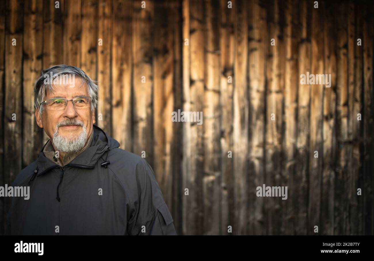 Uomo anziano che si gode all'aria aperta, escursioni, passeggiate attraverso la natura incantevole, in piedi di fronte a uno chalet di legno Foto Stock