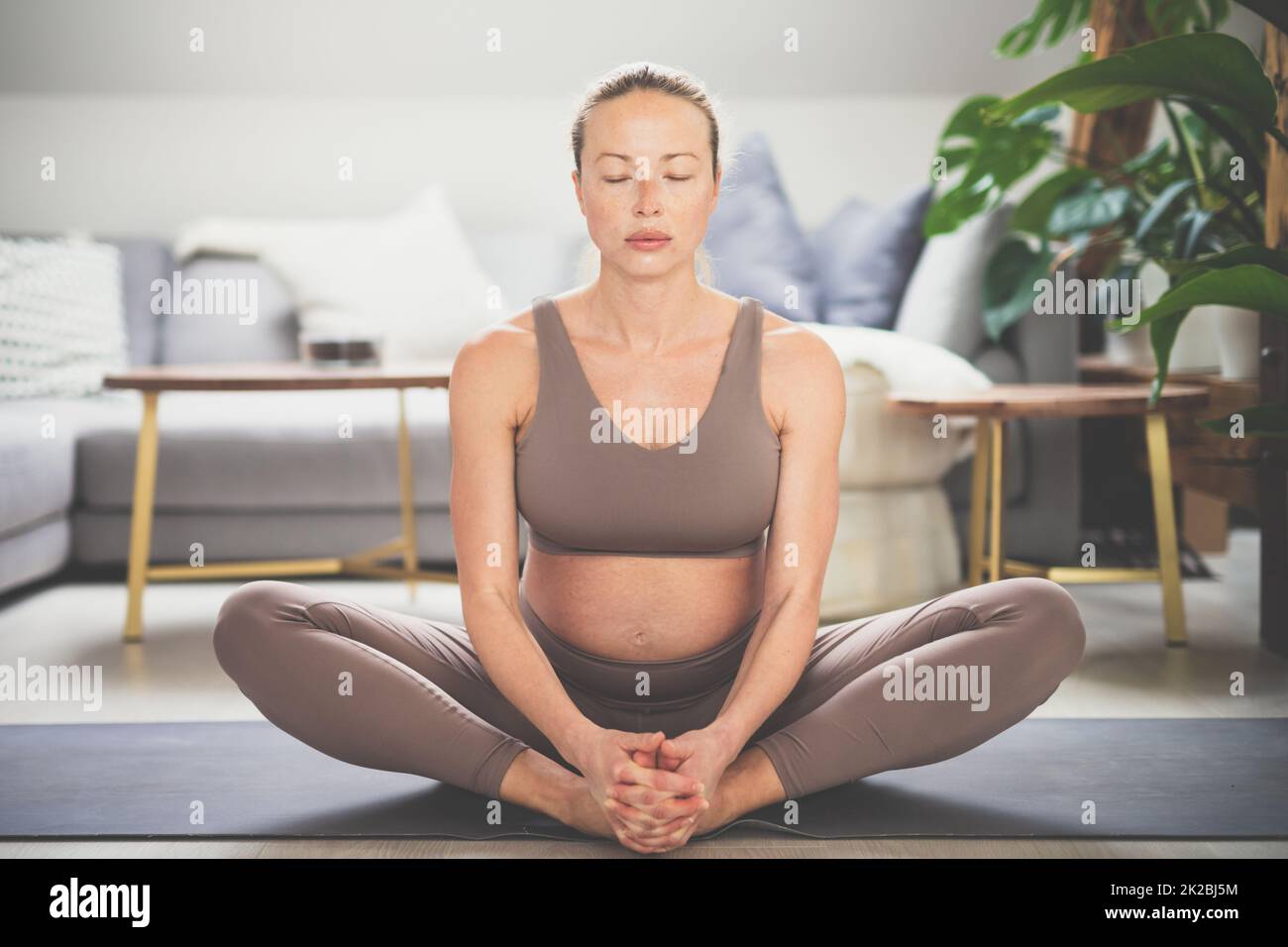 Giovane bella donna incinta che pratica yoga a casa nel suo soggiorno. Maternità, gravidanza, stile di vita sano, benessere e concetto di yoga. Foto Stock