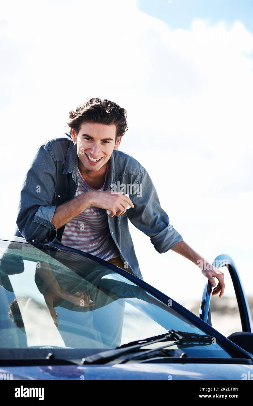 Pronto a colpire la strada. Felice uomo giovane che tiene un cellulare e si appoggia sul tetto della sua auto, sorridendo alla macchina fotografica. Foto Stock
