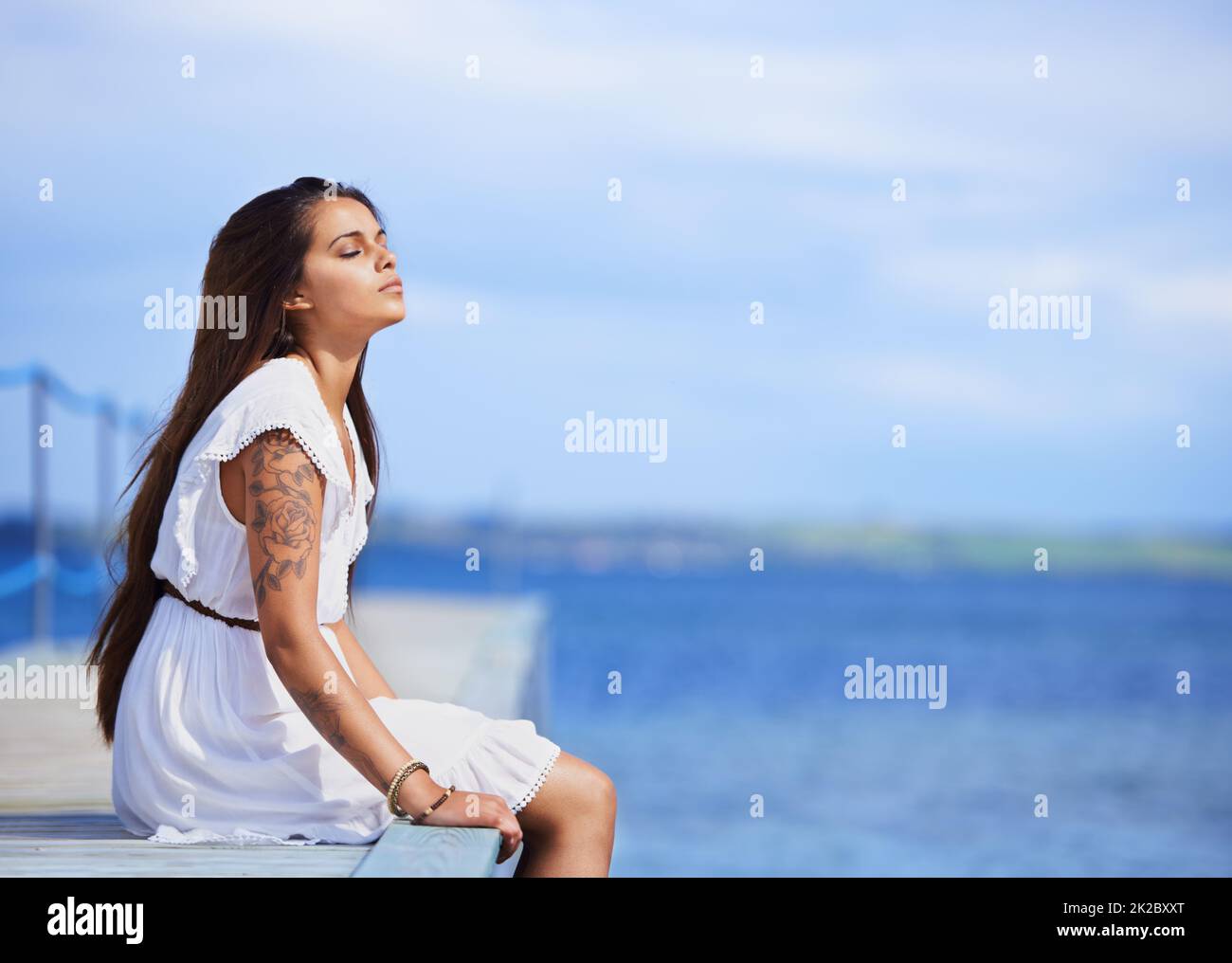 Contemplazione tranquilla accanto al mare. Scatto corto di una giovane donna bella tatuata seduta su un molo. Foto Stock