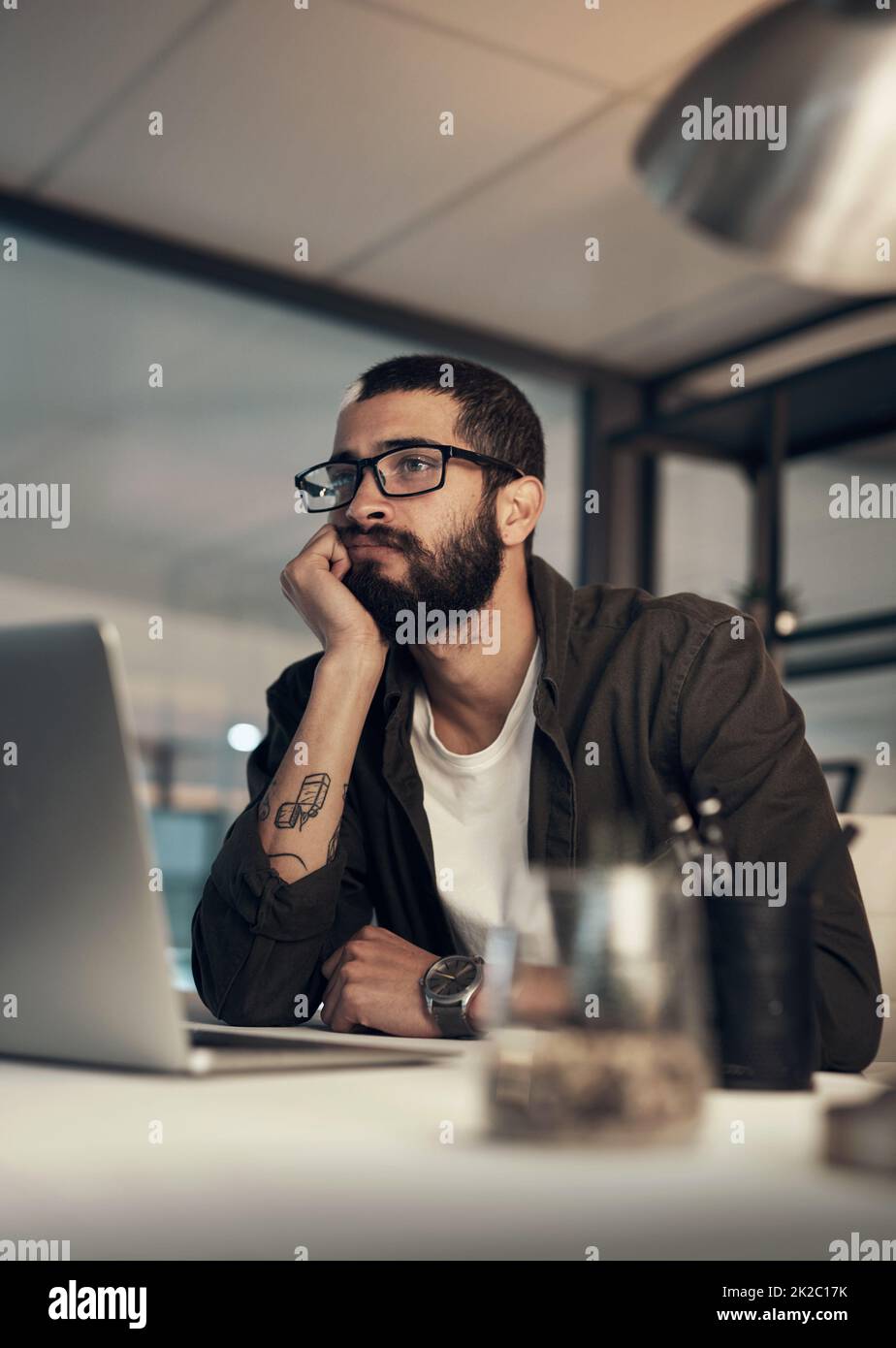 Meglio finire prima di addormentarmi. Scatto di un giovane uomo d'affari che guarda incerto mentre lavora a tarda notte in un ufficio moderno. Foto Stock