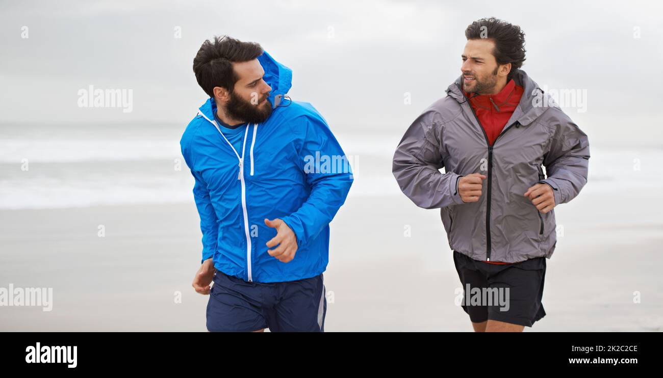 Una corsa mattutina sulla spiaggia. Scatto corto di due giovani uomini che corrono insieme lungo la spiaggia in una mattinata nuvolosa. Foto Stock