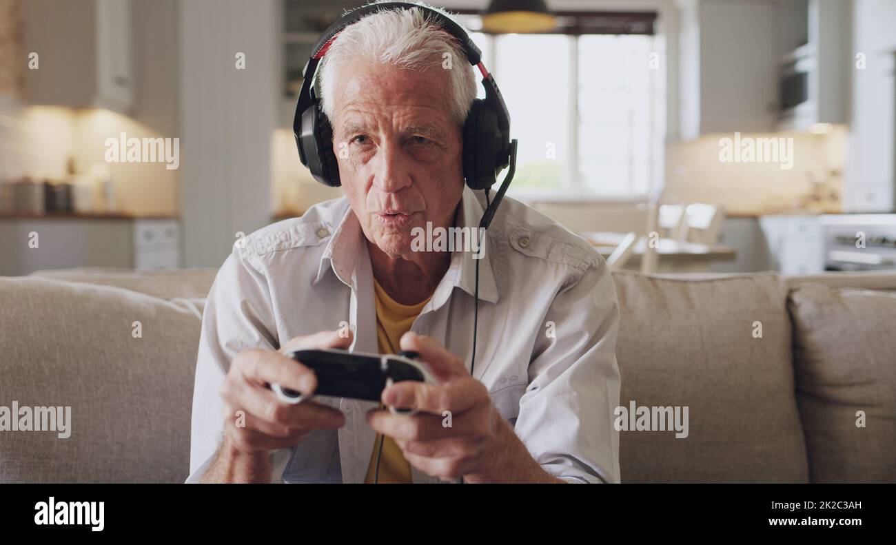 OOO, gli avversari che si avvicinano. Scatto di un uomo anziano seduto da solo sul divano a casa e indossando cuffie durante il gioco. Foto Stock
