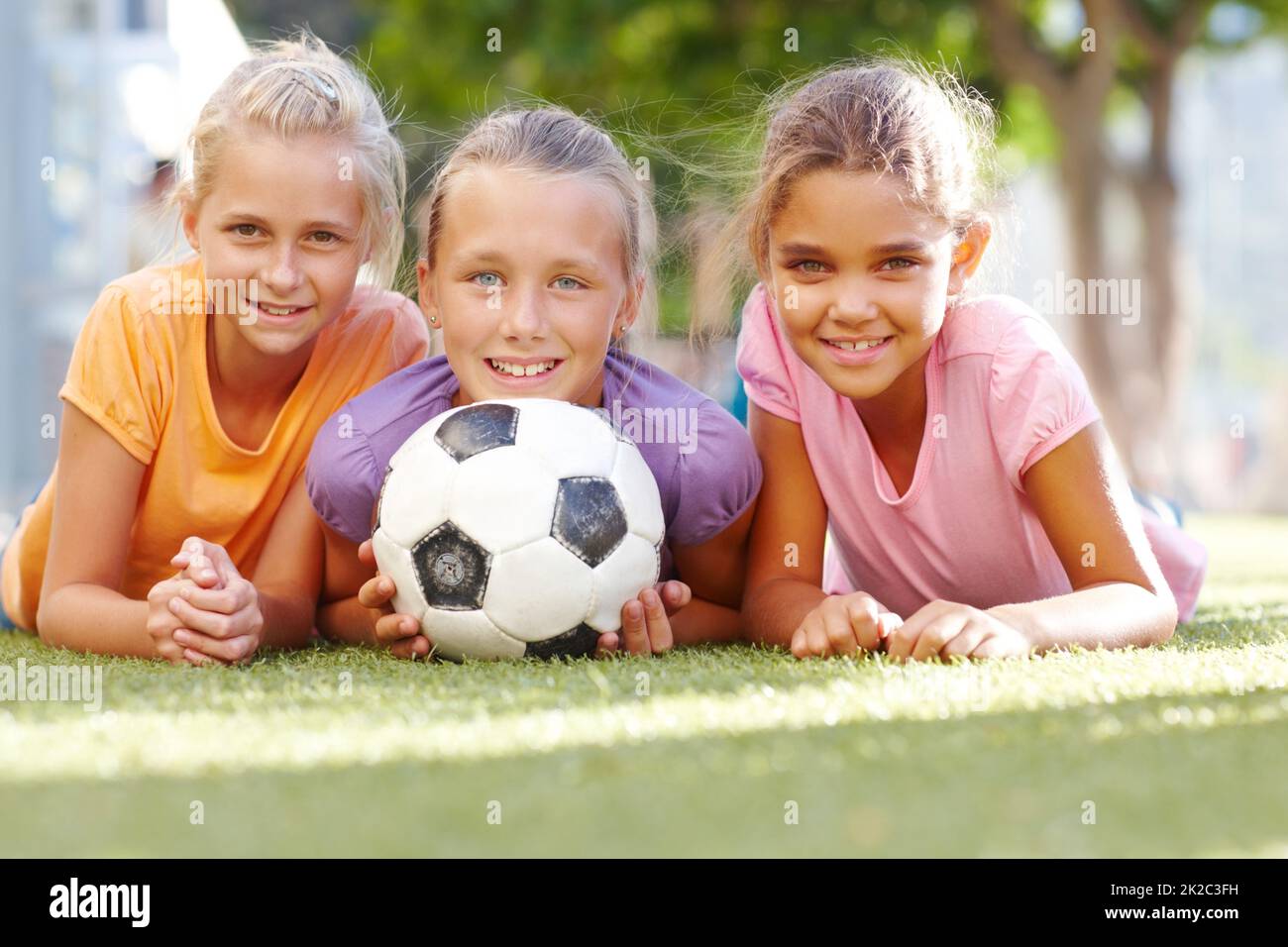 La nostra scuola offre calcio ragazze. Tre ragazze sorridenti che giacciono su un certo prato soleggiato che posano con una sfera di calcio copyspace. Foto Stock