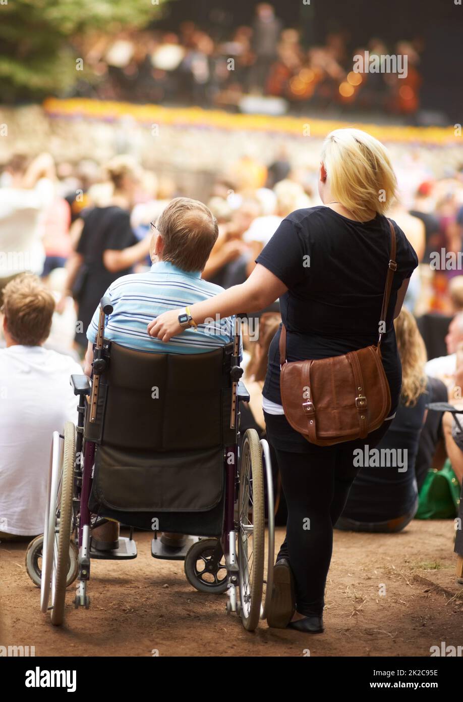 Il mio handicap non mi impedisce di godersi la vita. Vista posteriore di una moglie in piedi accanto al marito che è seduto in sedia a rotelle a un evento di musica dal vivo. Foto Stock