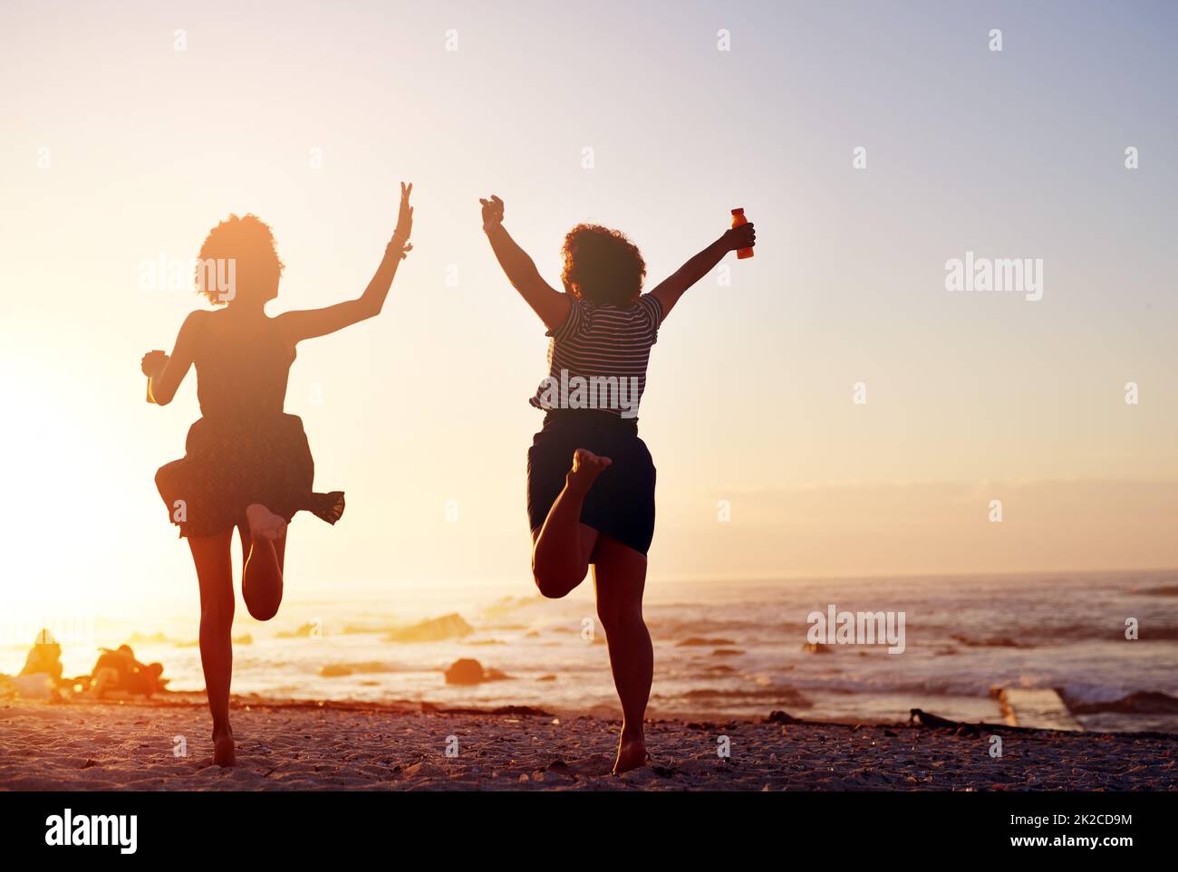 Amare la vita che viviamo insieme come amici. Ripresa da dietro di due giovani donne irriconoscibili che saltano nella gioia mentre camminano lungo la spiaggia al tramonto. Foto Stock