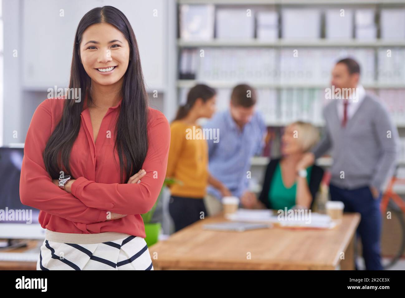Shell rendere il suo business un successo. Una giovane donna attraente in piedi con le braccia ripiegate mentre i suoi colleghi lavorano sullo sfondo. Foto Stock