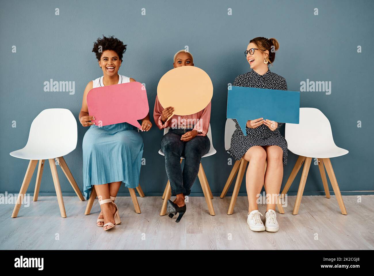 Credo di non avere detto questo. Studio shot di un gruppo di giovani donne d'affari attraenti che tengono bolle di discorso mentre si siedono in una fila contro uno sfondo grigio. Foto Stock