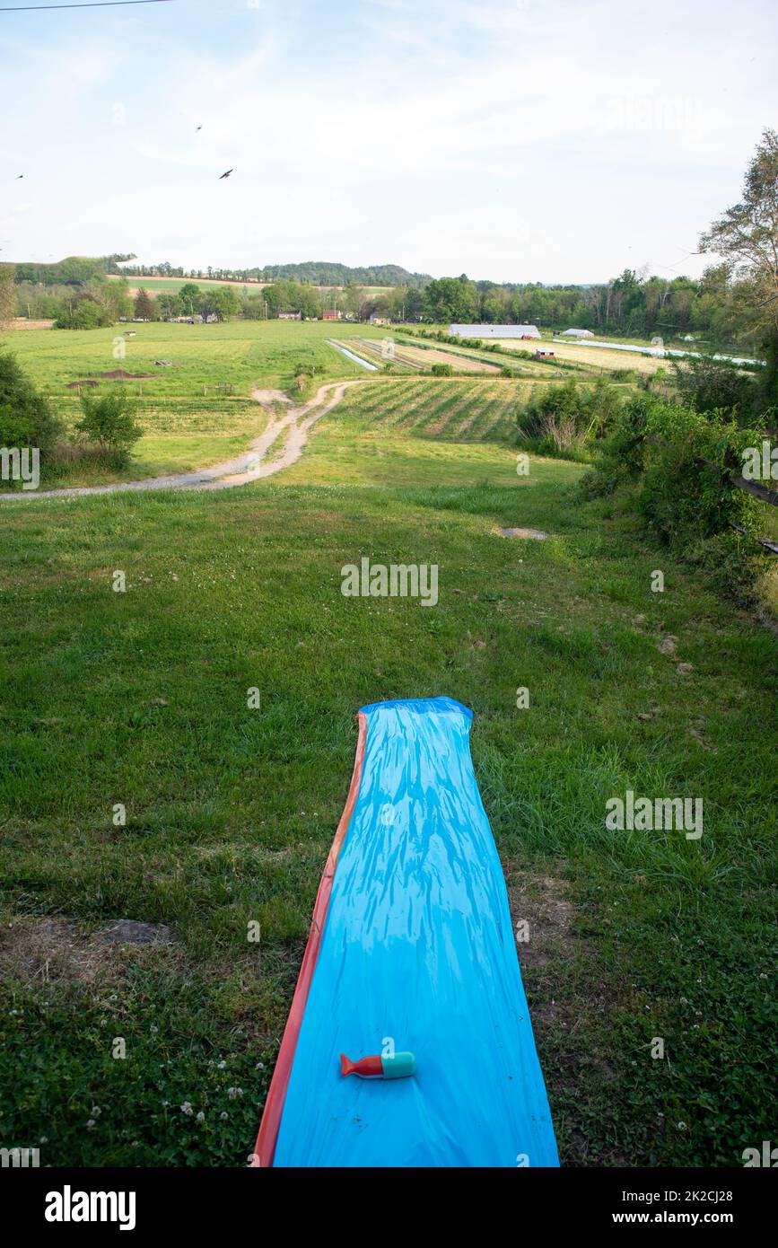 Scivolo d'acqua per bambini in plastica su una collina estiva erbosa sopra una fattoria Foto Stock