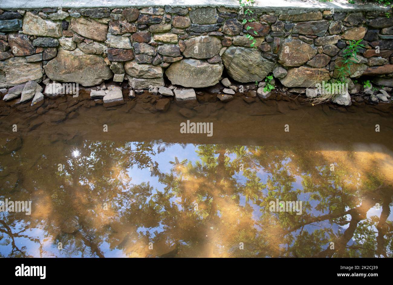 Muro di pietra con acqua calmo canale che riflette alberi e cielo Foto Stock