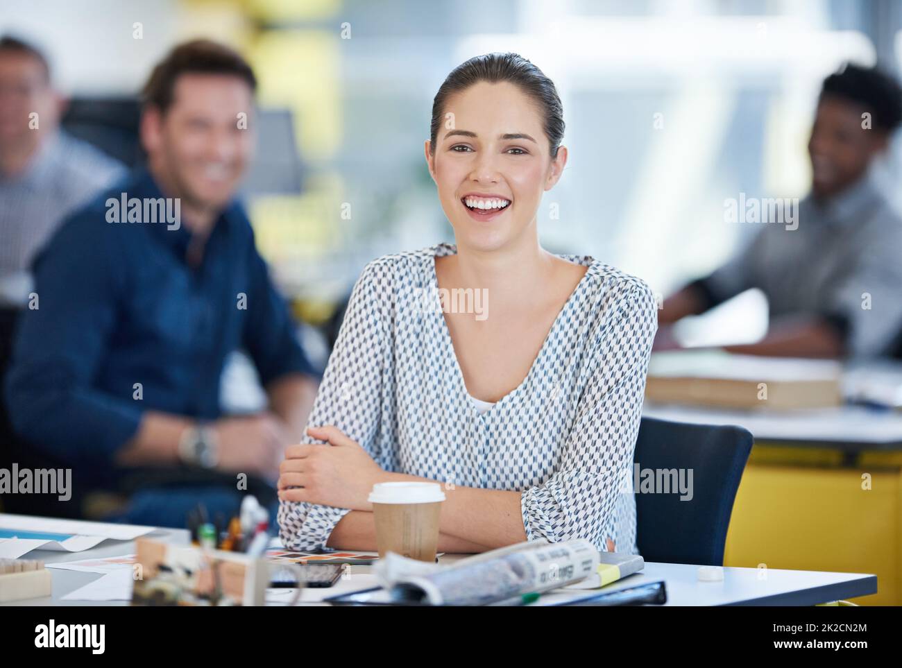 Non ho potuto chiedere un lavoro migliore. Ritratto di una giovane lavoratrice alla sua scrivania con i colleghi sullo sfondo. Foto Stock