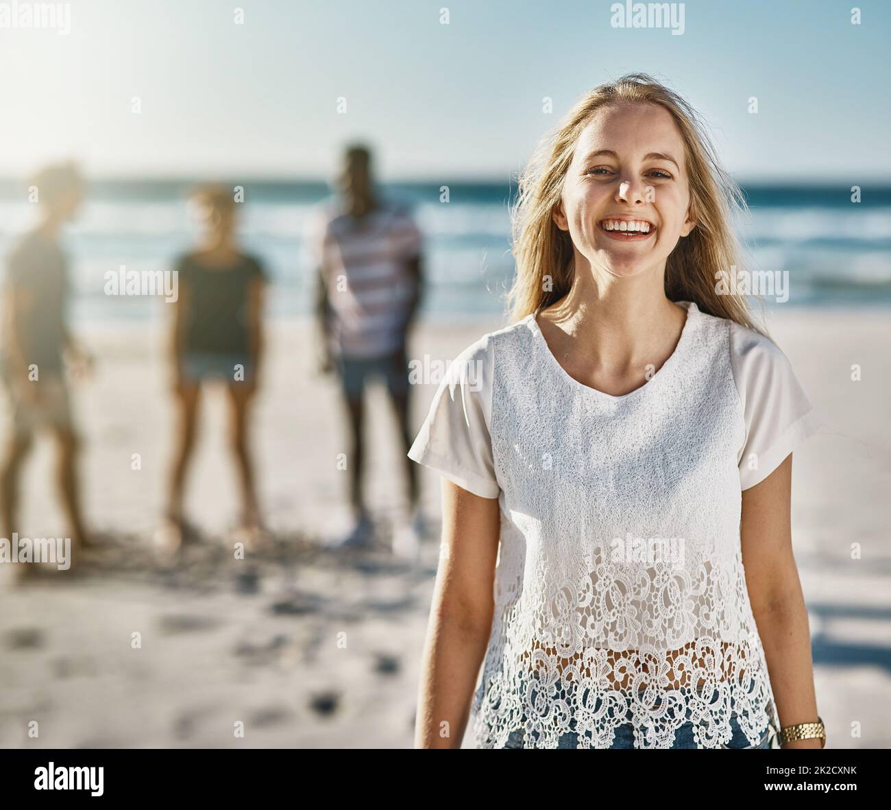 Non puoi fare a meno di sorridere quando il sole splende. Ritratto di una giovane donna felice in posa sulla spiaggia con i suoi amici sullo sfondo. Foto Stock