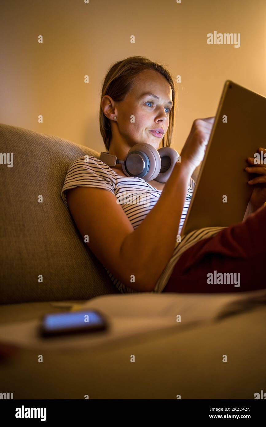 Piuttosto, giovane donna che lavorano a casa la sera - utilizzando il suo tablet pc come pure delle cuffie bluetooth, scrivere alcune idee per domani Foto Stock