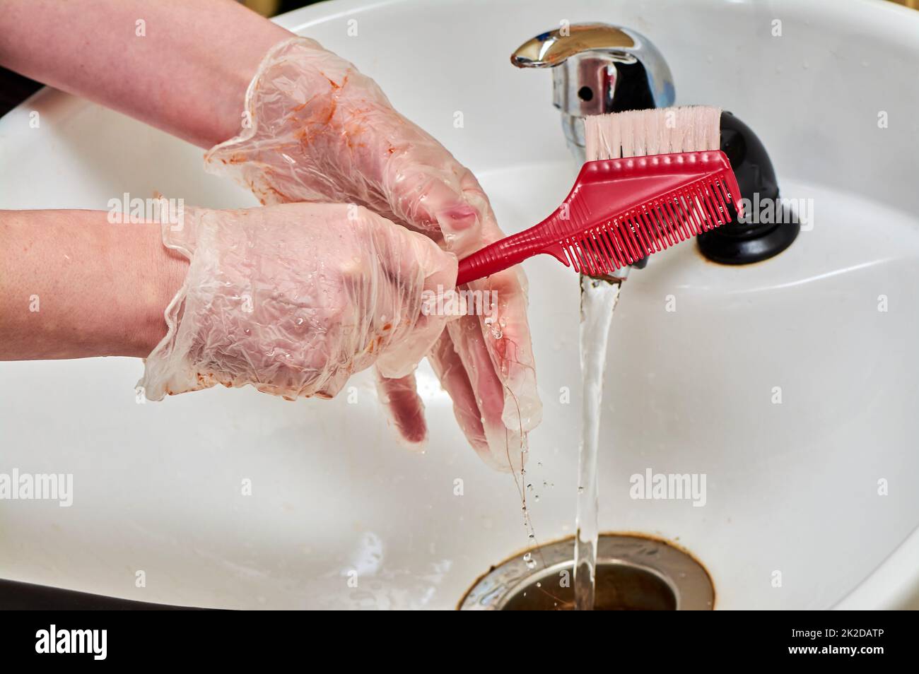 Le mani delle donne nei guanti da colorare dei capelli lavano un pettine sotto un flusso d'acqua in un lavandino bianco Foto Stock
