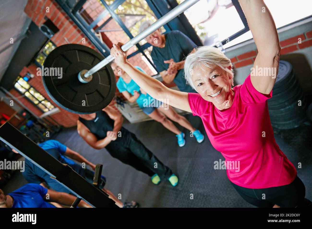 Non diventa più facile, si ottiene più forte. Colpo di una donna anziana sollevamento pesi mentre un gruppo di persone in background guardare sopra. Foto Stock