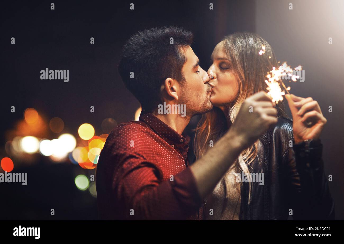 Le scintille volano quando baciamo. Scatto di una giovane coppia felice che festeggia con i luccicanti all'aperto di notte. Foto Stock
