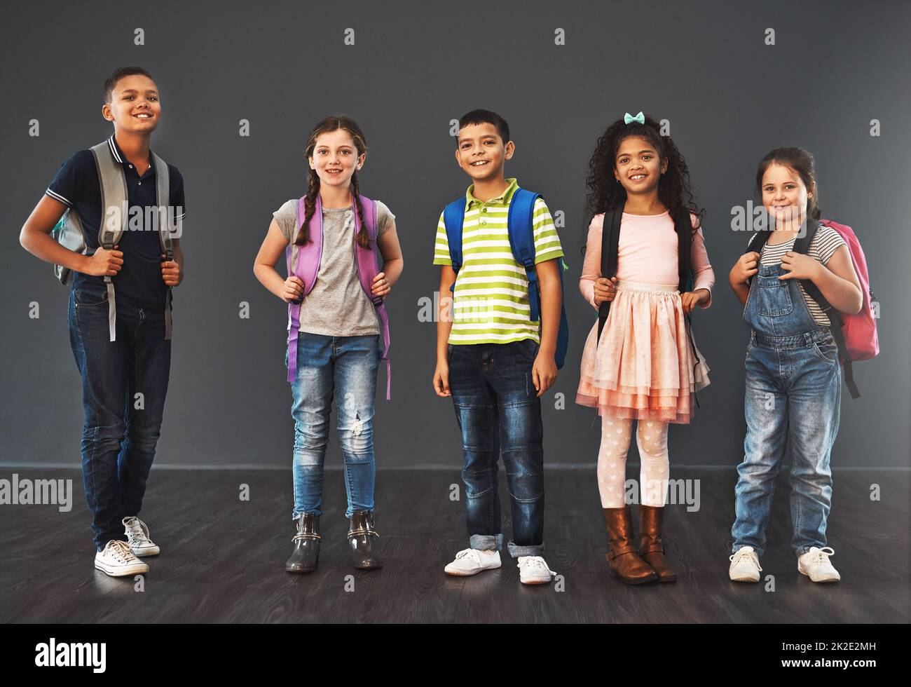 Gente piccola, grande giorno. Ritratto di studio di un gruppo diversificato di bambini che trasportano i loro zaini della scuola su uno sfondo grigio. Foto Stock