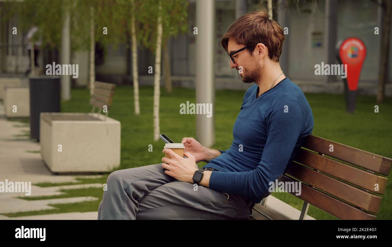 Scena all'aperto di un bel giovane uomo che naviga nell'app social network per smartphone mentre beve un caffè. Studente uomo rilassante in un parco. Foto Stock