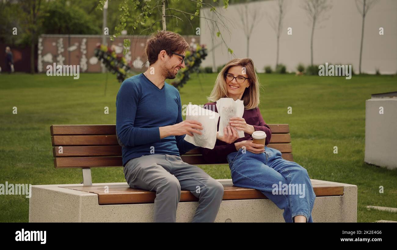 Un bell'uomo porta il pranzo per un collega seduto su una panca nel parco. Pausa caffè di lavoro. Tempo di relax per studenti. Passeggiata all'aperto. Concetto di cura. Foto Stock