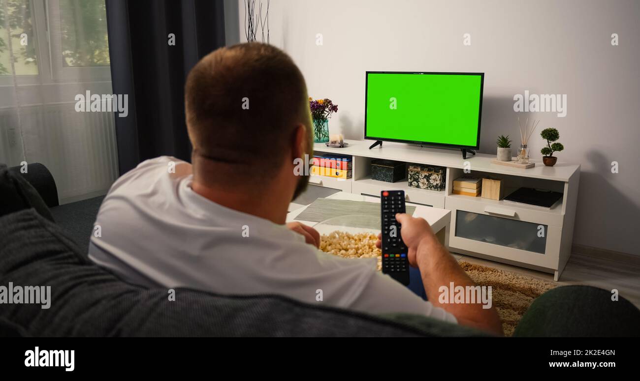 Vista posteriore uomo seduto sul divano nel soggiorno guardando la TV con schermo verde e cambiando canali con telecomando in mano. Chroma tasto TV in messa a fuoco. Foto Stock