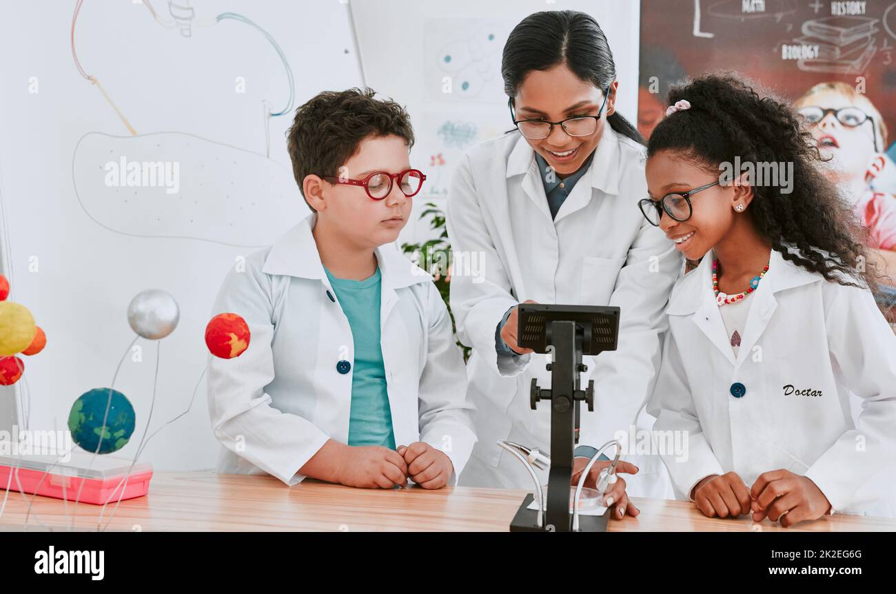 Diamo un'occhiata più da vicino a questo. Shot di una giovane insegnante di scienza usando un microscopio con le sue alunne in classe di scienza a scuola. Foto Stock