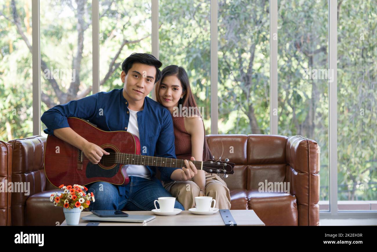 I giovani amanti trascorrono del tempo insieme in vacanza nel soggiorno. Il giovane indossa abiti comodi, suona una chitarra. Foto Stock