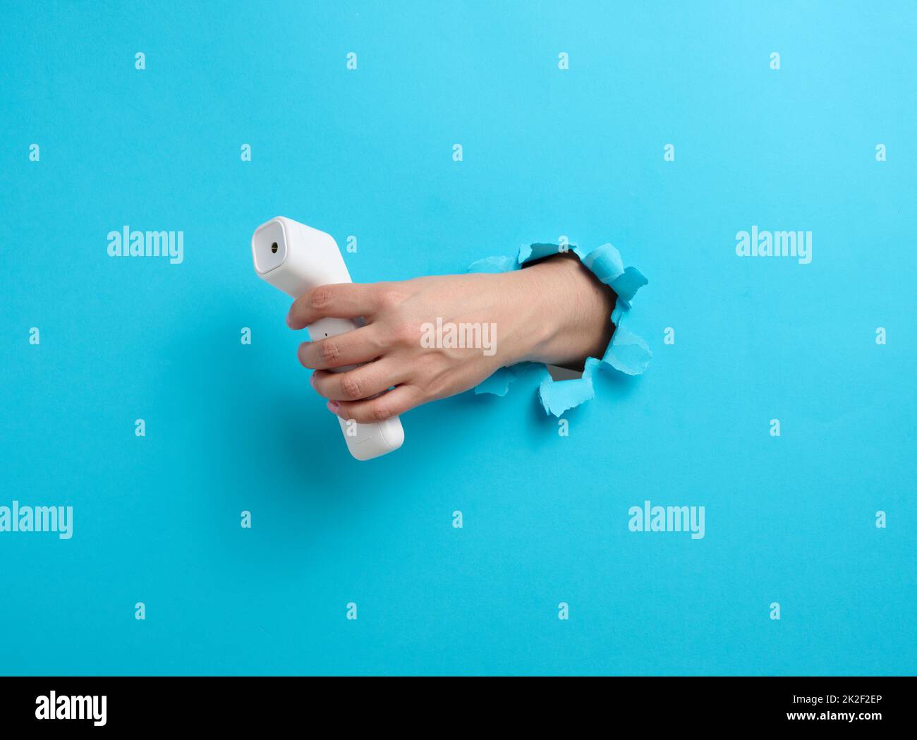 una mano femmina tiene un termometro in plastica senza contatto per misurare la temperatura di una persona, una parte del corpo sporge da un foro lacerato su uno sfondo di carta blu Foto Stock