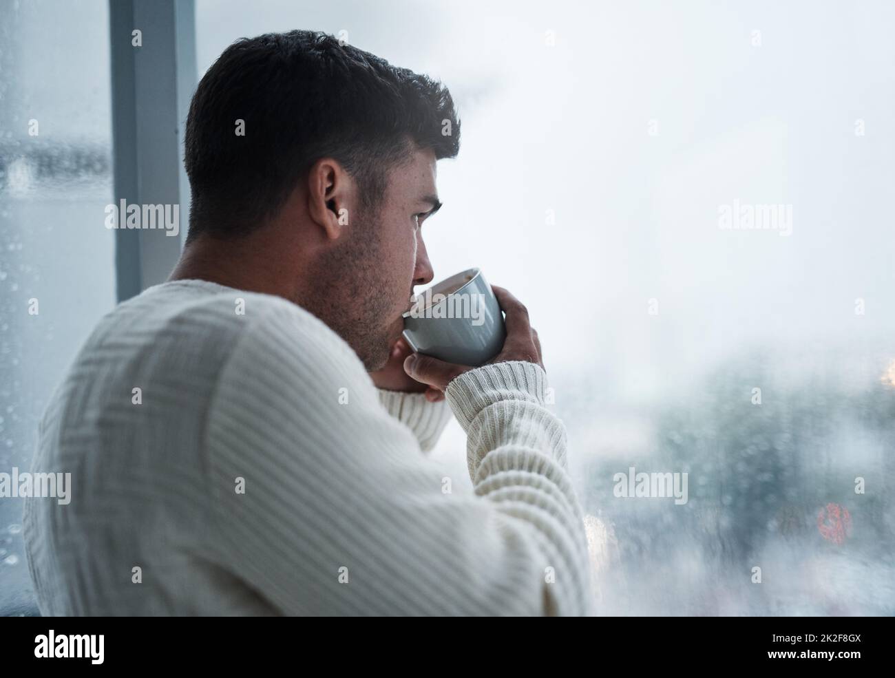 Una semplice pausa vi riporterà a voi. Scatto di un giovane uomo che ha il caffè e guardando fuori da una finestra in una giornata piovosa a casa. Foto Stock