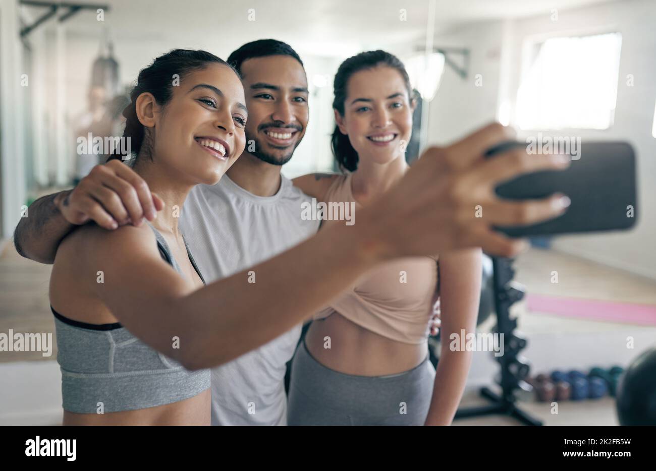 Online per diffondere un po' di ispirazione per il fitness. Scatto di tre giovani atleti che prendono un selfie mentre si levano in piedi insieme nella palestra. Foto Stock
