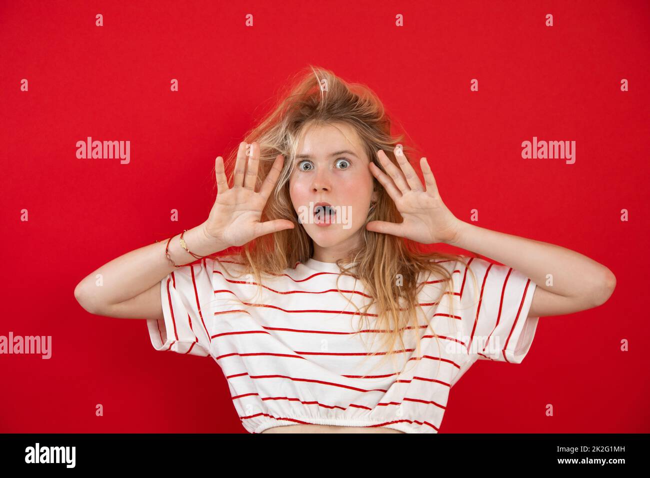 Stativo di giovane signora con gli occhi aperti con le mani sollevate con le barrette sparse vicino al viso su fondo rosso vuoto. Bella ragazza con capelli selvatici hanno divertimento e spettacolo Foto Stock