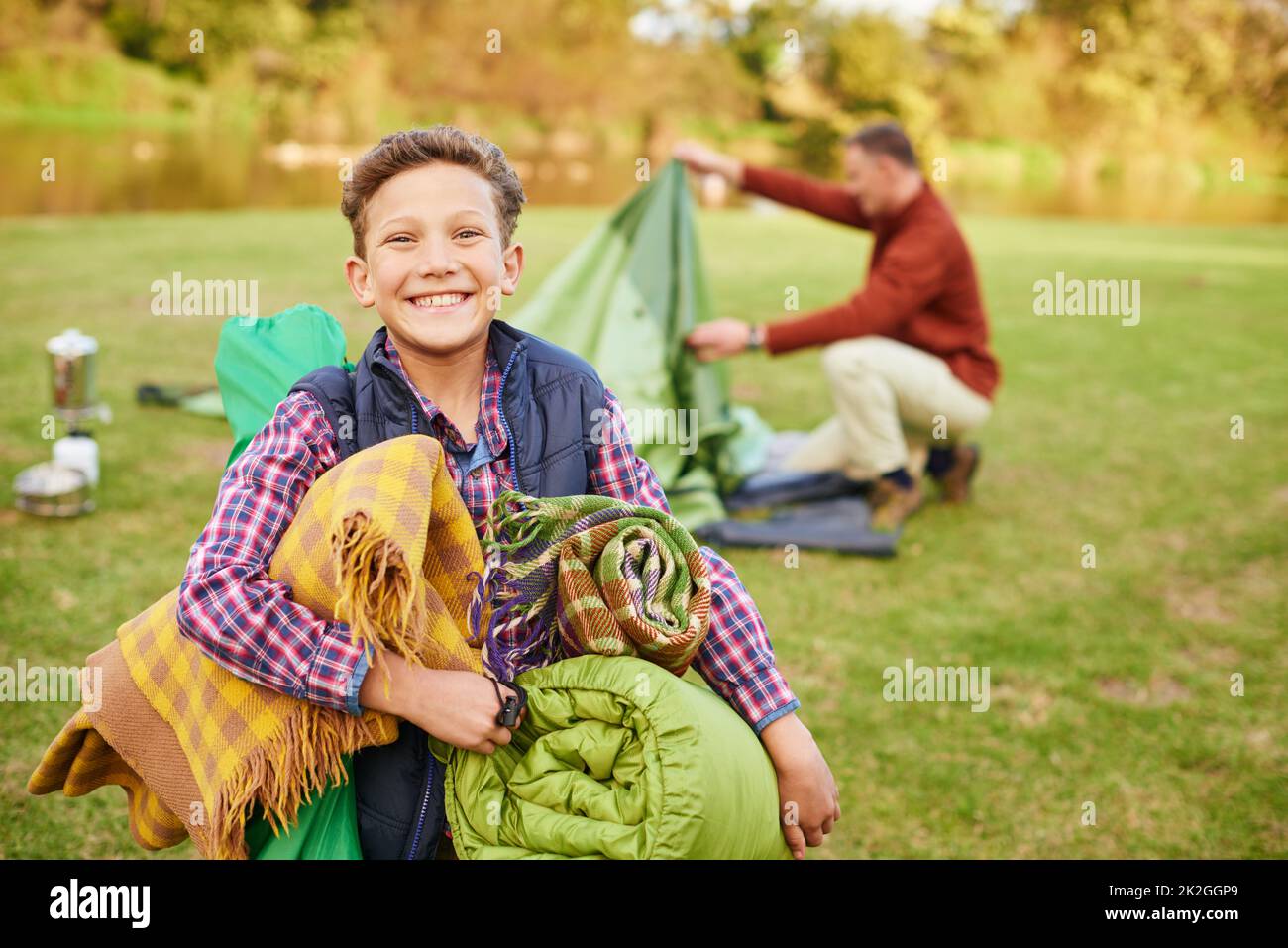 Questo viaggio in campeggio sarà impressionante. Ritratto di un ragazzo che tiene l'attrezzatura da campeggio con il padre sullo sfondo. Foto Stock