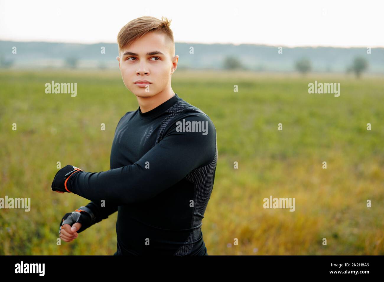 Primo piano ritratto, giovane uomo in camicia nera, stendendo le braccia, isolato verde sfondo alberi. Riscaldamento in corso Foto Stock