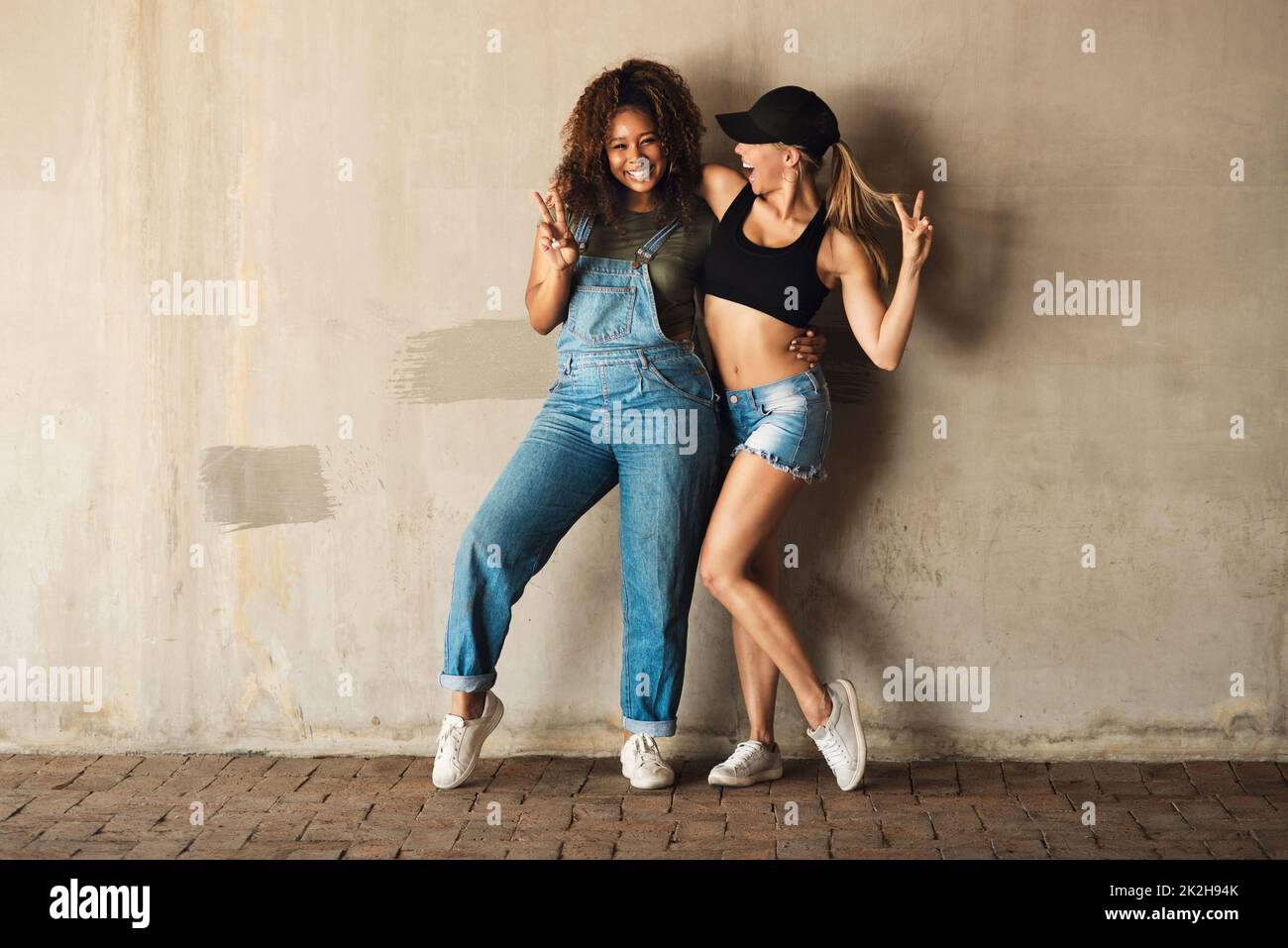 Sappiamo come Buon divertimento. Ritratto di due giovani donne allegre in posa per una foto mentre si appoggia contro una parete esterna durante il giorno. Foto Stock