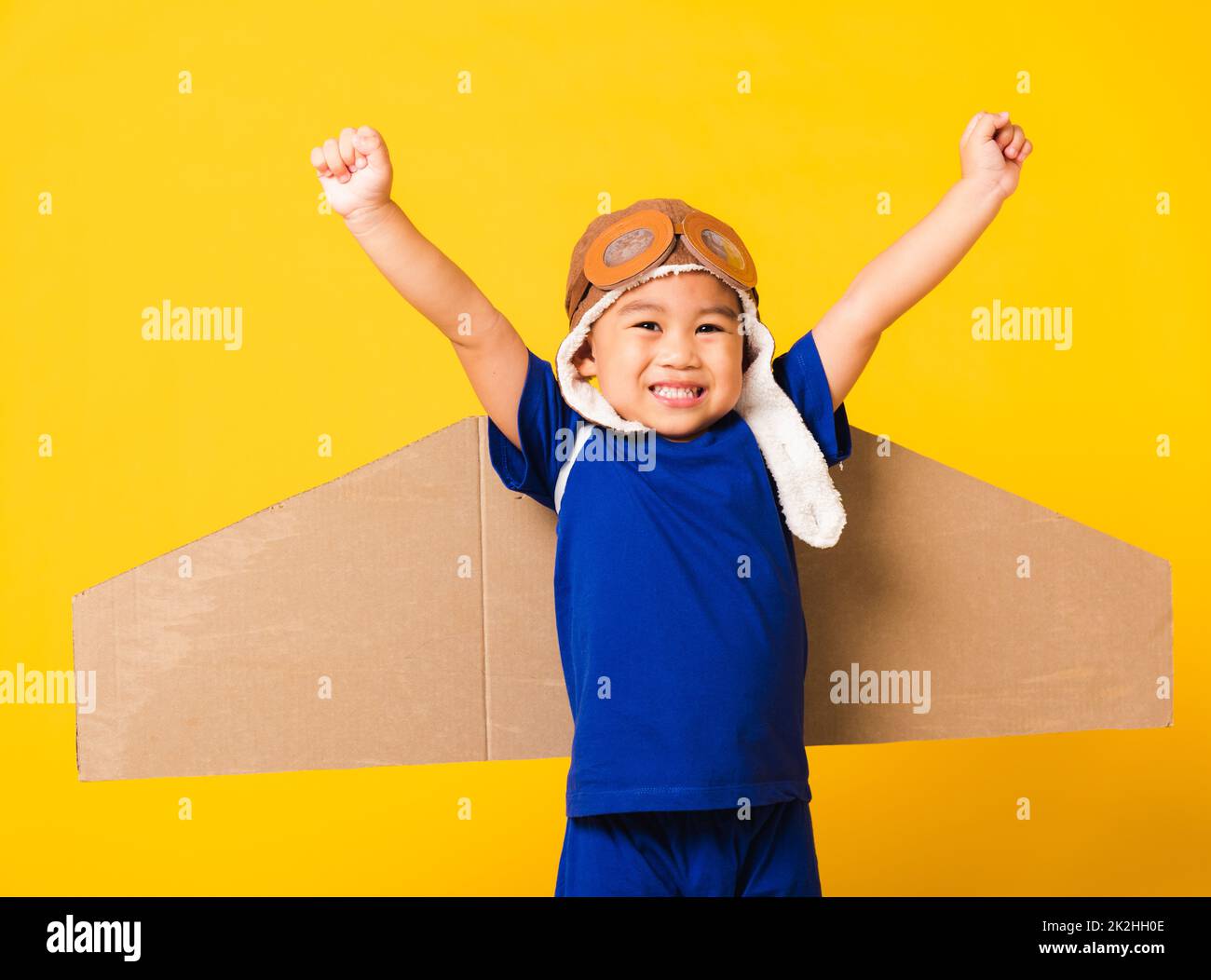 Bambino piccolo ragazzo sorriso indossare pilota cappello gioco e occhiali di protezione con ali di aeroplano giocattolo in cartone Foto Stock