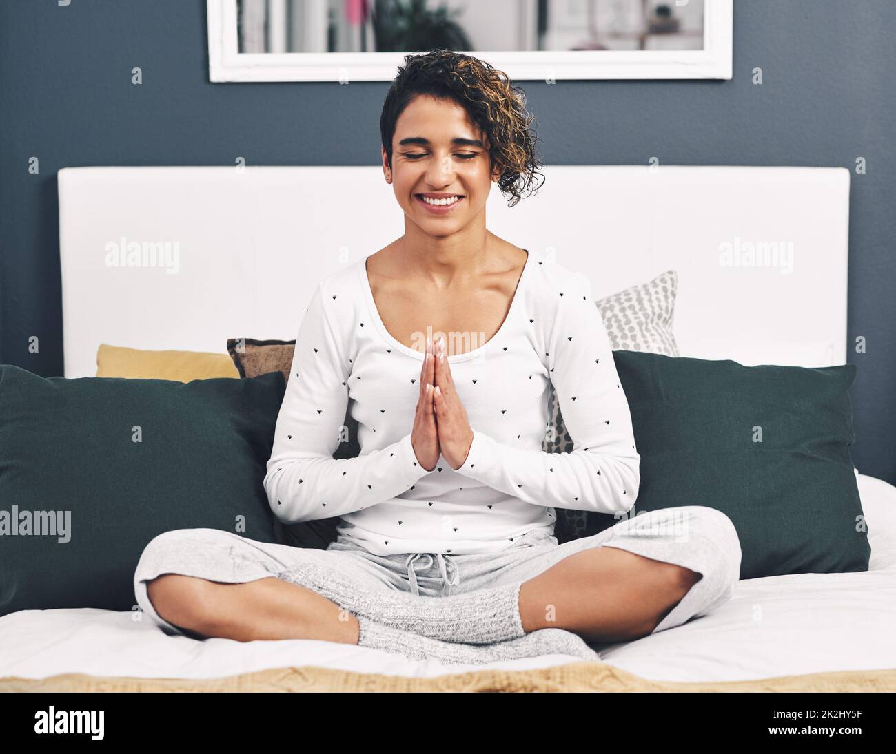 La meditazione ha cambiato la mia vita. Scatto a tutta lunghezza di una giovane donna sedente attraente a croce legato sul suo letto a casa con le sue palme insieme. Foto Stock